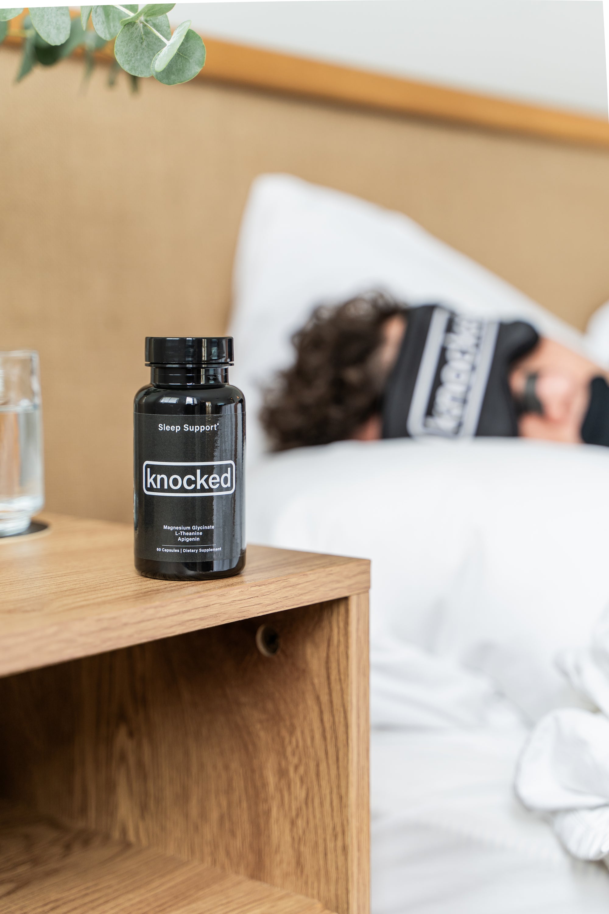 Knocked sleep support on the bedside table. In the background a man sleeping in bed with knocked eyemask, nose strips, and mouth tape.